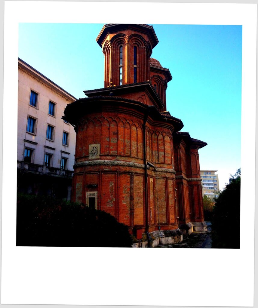 Polaroid photograph of a Romanian orthodox church in red brick with an octagonal tower and decorative arches in a yellowish-white style, framed between modern white buildings, against an intense blue sky.