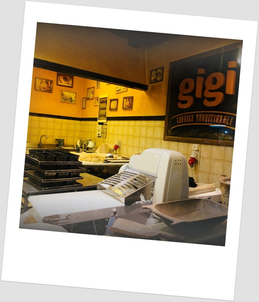 Fotografia polaroid do interior de uma padaria tradicional romena "Gigi Covrigi Tradiționali" em Bucareste, com máquina de cortar pão, tabuleiros negros empilhados, balcão de preparação com massa e parede amarela com fotografias emolduradas.
