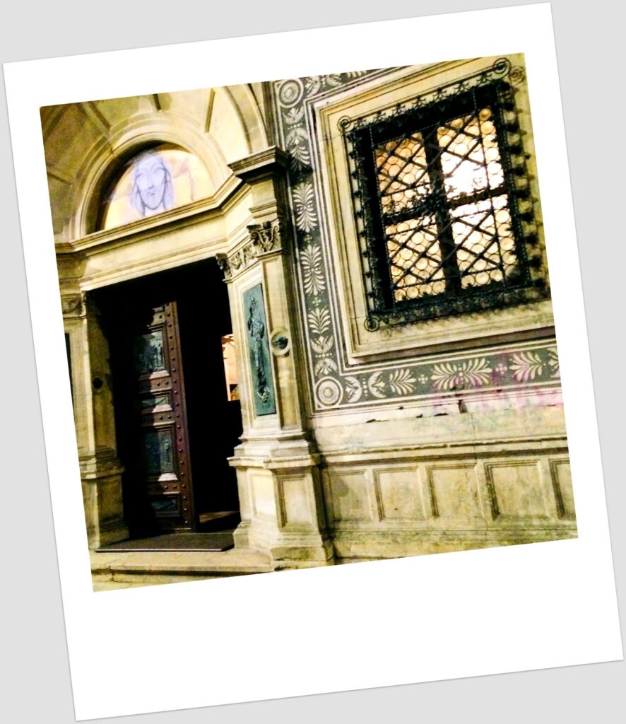 Fotografia polaroid do portal lateral do Ateneu Romeno em Bucareste, com porta de madeira escura entalhada, afresco de rosto feminino em arco semicircular, janela com gradeamento de ferro forjado e paredes com frisos florais pintados a verde e branco.