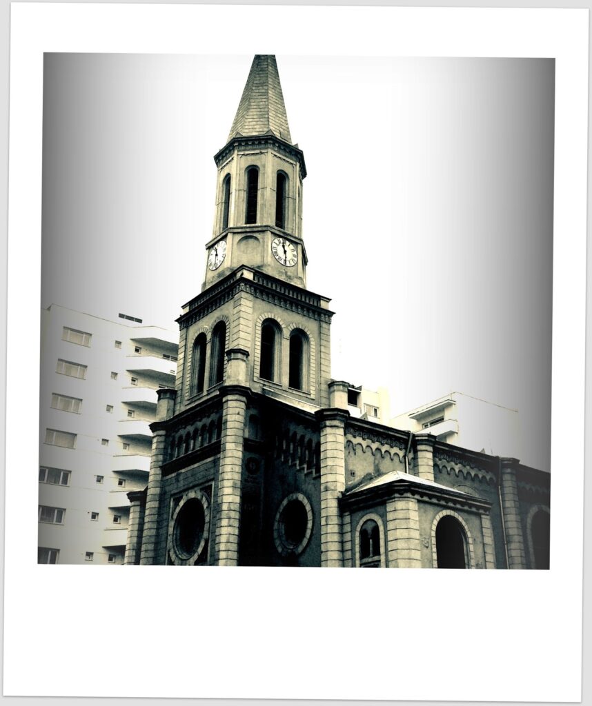 Fotografia polaroid em tons a preto e branco de uma igreja com campanário octogonal e agulha pontiaguda com dois relógios de algarismos romanos, rodeada por blocos de apartamentos brancos, em céu encoberto.