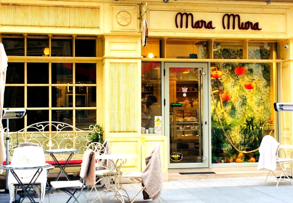 Fachada exterior da pastelaria e café "Mara Mura" em Bucareste, com insígnia em letras cursivas escuras, fachada amarela com grandes janelas, esplanada com mesas e cadeiras de ferro forjado branco com mantas e decoração tropical com lanternas vermelhas visível no interior.