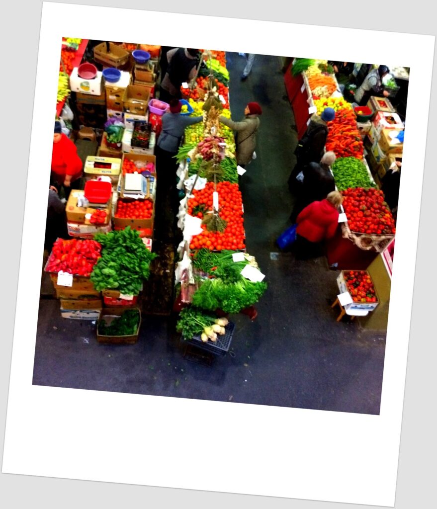 Vista aérea de mercado coberto de frutas e legumes em Bucareste com bancas coloridas de tomates, ervas verdes, cenouras, alfaces e outros produtos, com clientes a fazer compras entre os corredores.