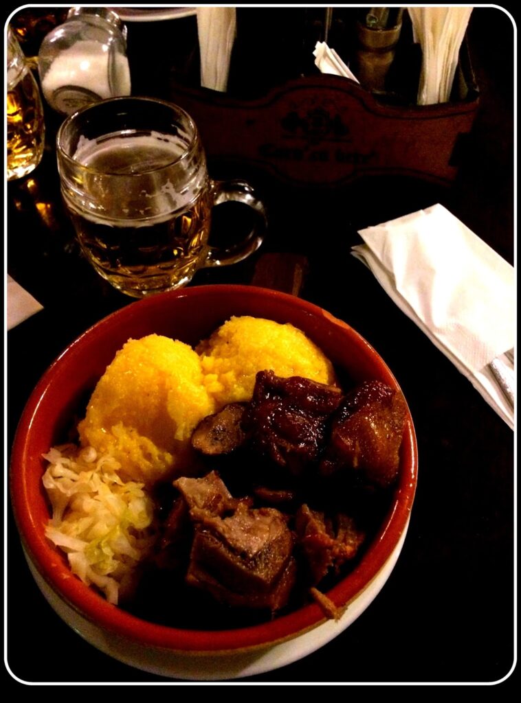 Red clay bowl with mămăligă (polenta) in yellow, pieces of roast pork and varză acră (Romanian sauerkraut), on a dark restaurant table, with a faceted glass beer mug in the background.