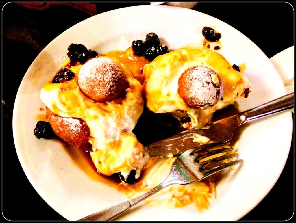 Fotografia polaroid de prato branco com dois papanași (donuts romenos fritos) cobertos com natas brancas e compota de frutos negros, com bola de massa pequena no topo de cada um, em mesa escura de restaurante.