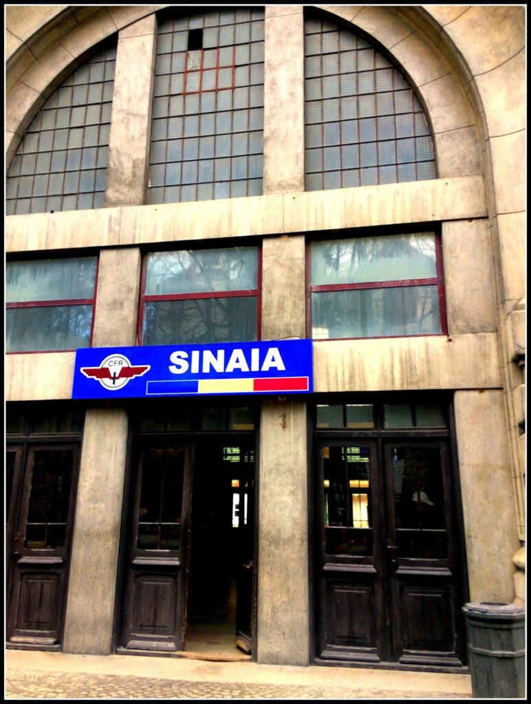 Fotografia polaroid da fachada da estação de comboios de Sinaia, com grande janela arqueada em ferro e vidro, painel azul com inscrição "SINAIA" e logótipo CFR com bandeira romena, portas de madeira escura e pavimento de calçada.