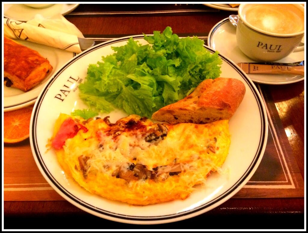 White plate with Paul logo with an open mushroom and cheese omelette, green lettuce salad, baguette slice, half an orange, on a wooden tray with a cappuccino in a Paul cup, croissant and napkin alongside.