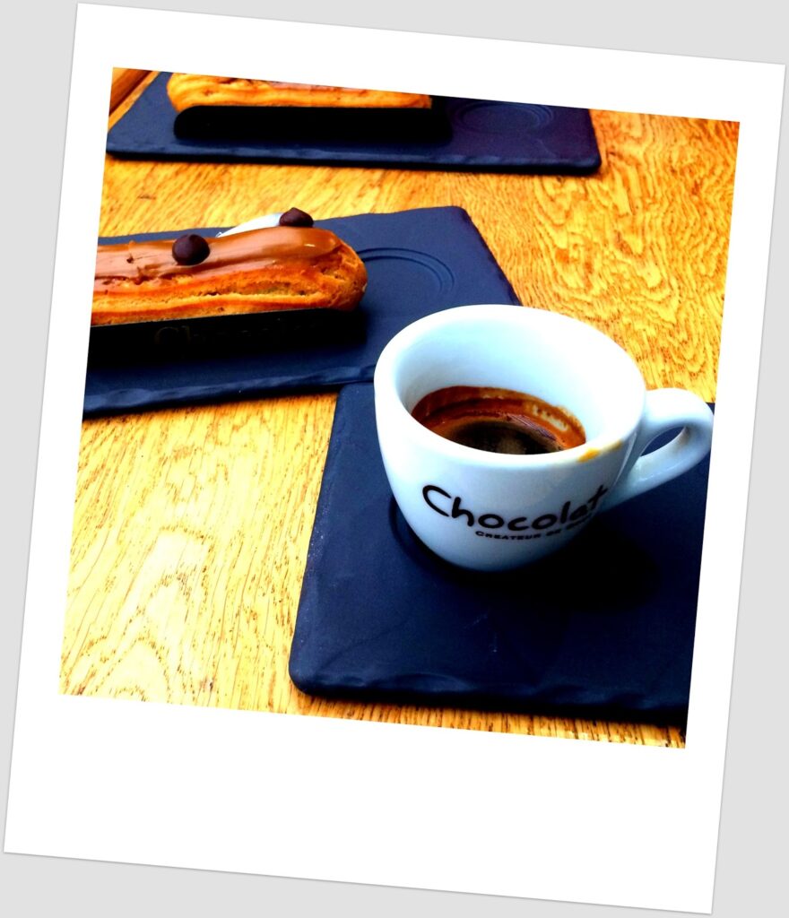 Tilted polaroid photograph of two black slate trays with chocolate éclairs with glossy coating and a chocolate chip, and a white espresso cup inscribed "Chocolat" on a slate tray, on a light wooden table.