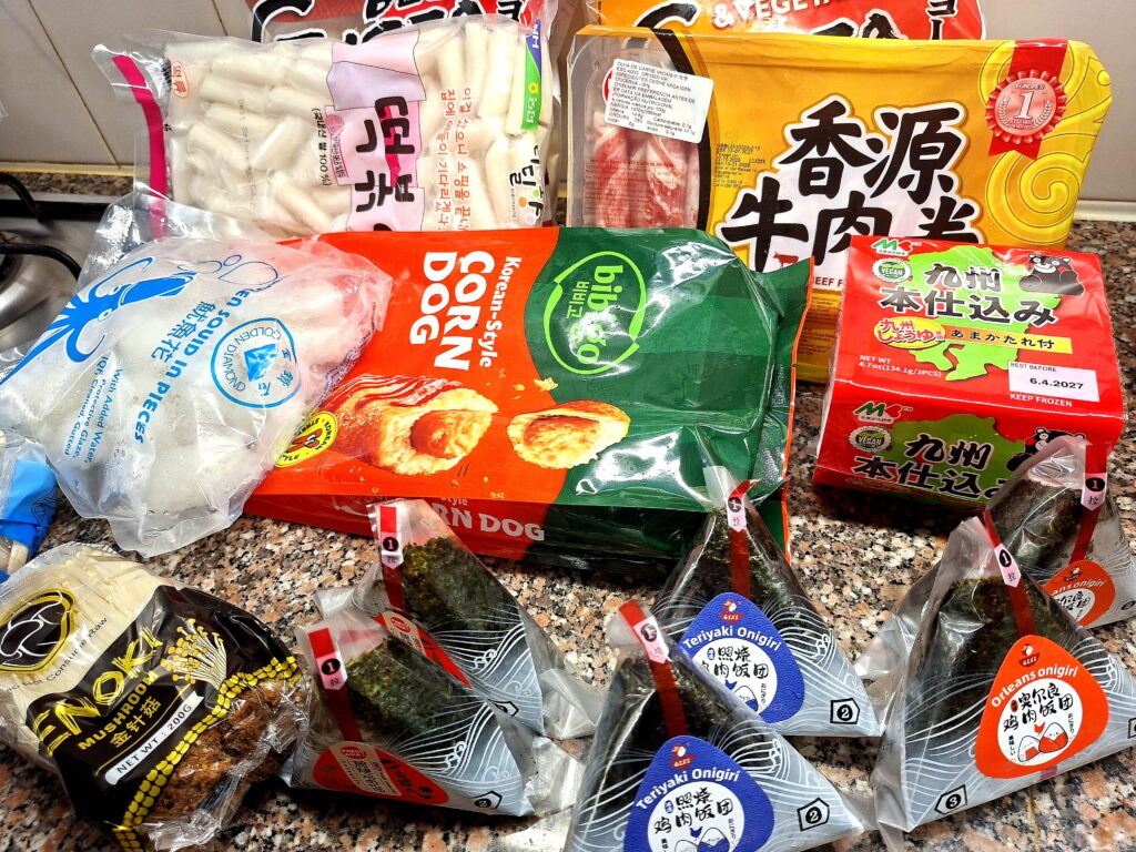 Kitchen counter with various frozen and packaged Asian food products: Ajinomoto meat and chicken gyoza, Korean tteok, frozen squid, Korean corn dogs, rice balls, sliced beef, Japanese ramen, enoki mushrooms, and individually wrapped teriyaki onigiri.