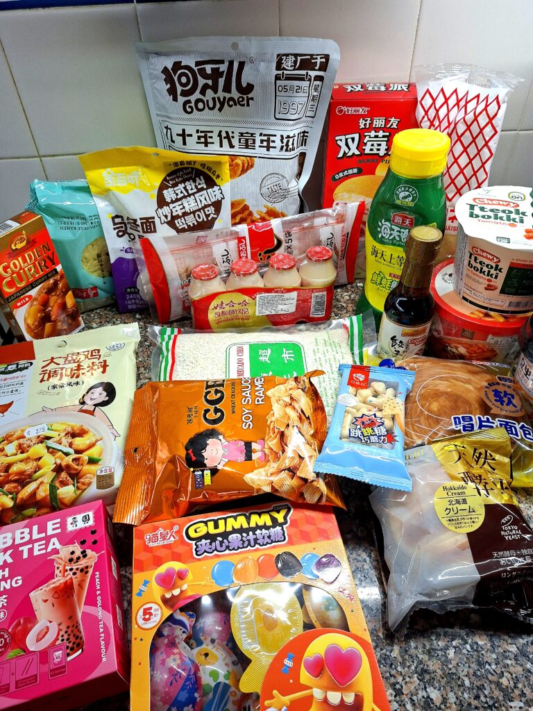 Kitchen counter with a wide variety of dry and packaged Asian food products: Japanese Golden Curry, ramen, chicken seasoning, peach bubble tea, Gouyaer cookies, mini Wahaha yogurts, soy sauce, instant tteokbokki, rice, wheat crackers, gummies, Hokkaido bread, and other snacks with colorful packaging in Chinese, Japanese, and English.
