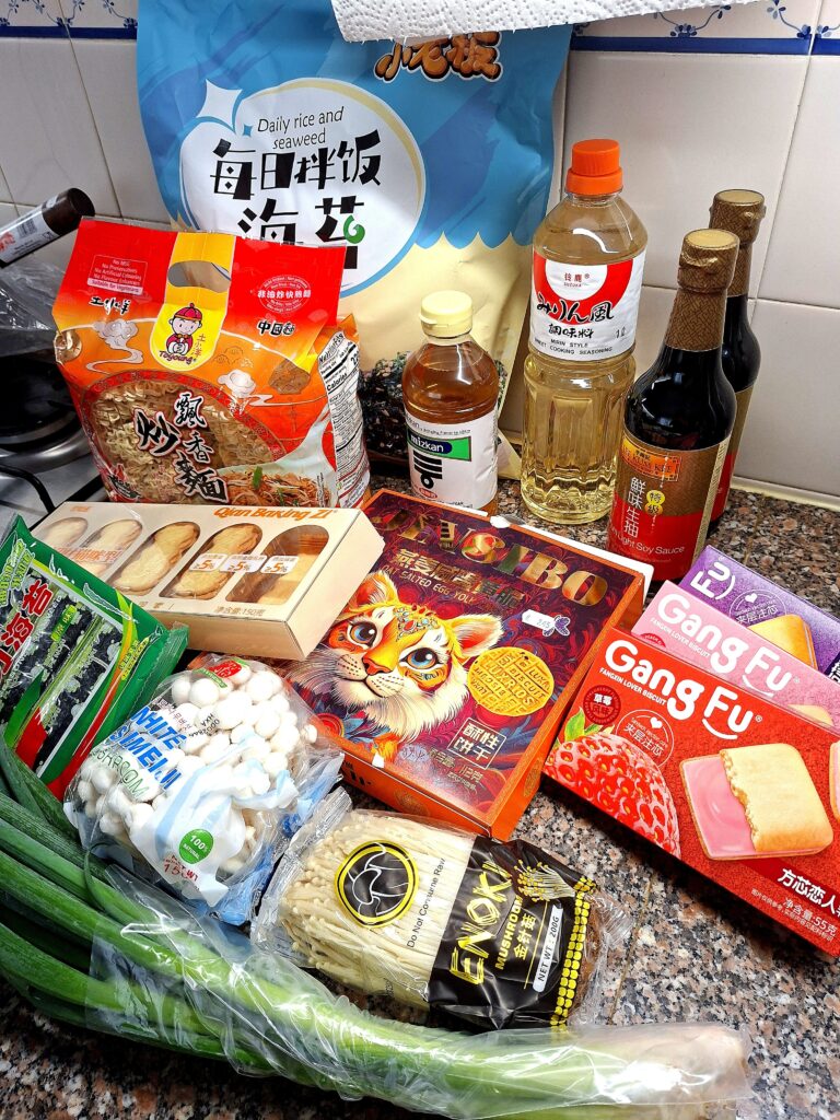 Bancada de cozinha com produtos asiáticos: saco grande de algas para arroz diário, dois pacotes de noodles fritos chineses, garrafas de mizkan, mirin e molho de soja light, caixa de salted egg yolk biscoitos, biscuits Gang Fu de morango, caixas de biscoitos japoneses, enoki, cogumelos shimeji brancos, cebolinho fresco e cebola.