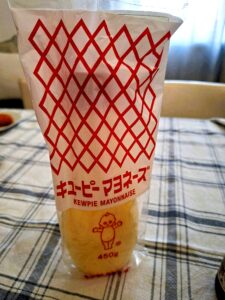 Embalagem frontal da Kewpie Mayonnaise japonesa de 450g, com padrão xadrez vermelho e branco, logótipo em japonês e inglês e a boneca Kewpie desenhada a vermelho, pousada sobre toalha de mesa às riscas azuis e brancas.