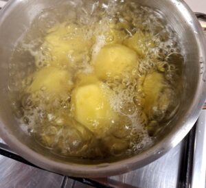 Aerial view of a stainless steel pot with whole peeled potatoes boiling vigorously in water, on a stainless steel gas hob, with white tiles in the background.