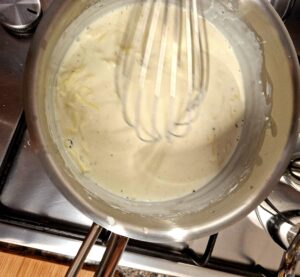 Top-down view of a stainless steel pan on the hob with white and creamy béchamel sauce being whisked with a metal balloon whisk, with another pan visible in the background.
