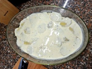 Vista aérea de travessa oval de vidro com batatas fatiadas cobertas com molho béchamel branco e cremoso, sobre bancada de granito, com faca e tábua de madeira Tefal visíveis ao lado.