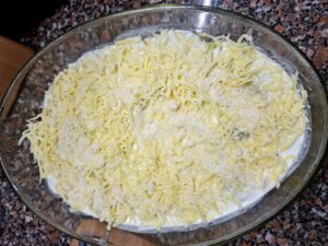 Vista aérea de travessa oval de vidro com batatas e béchamel cobertos por uma camada generosa de queijo ralado amarelo-pálido misturado com pão ralado, sobre bancada de granito.