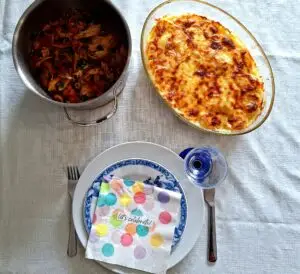 Vista aérea de mesa posta com panela de frango estufado desfiado com cenoura e ervas, travessa oval de gratinado de batata dourado, prato de faiança azul e branca com guardanapo colorido "let's celebrate", copo azul e talheres.
