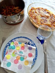 Table with a white linen cloth with a stainless steel pot of braised chicken on the left, an oval glass baking dish with golden potato gratin on the right, a blue and white faience plate with a festive "let's celebrate" napkin, a blue-stemmed glass and cutlery.