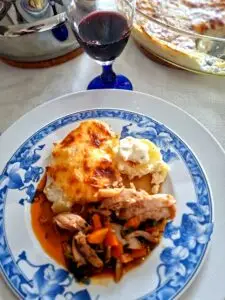 Prato de faiança azul e branca com porção de frango estufado em molho alaranjado com cenoura e ervas, acompanhado de porção de gratinado de batata dourado e creme branco, com copo de vinho tinto em primeiro plano e travessa ao fundo.
