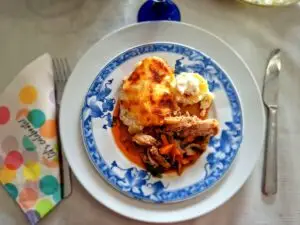 Aerial view of a blue and white faience plate with a portion of toasted potato gratin, white cream and shredded braised chicken with carrot in an orange sauce, flanked by a festive napkin and cutlery.