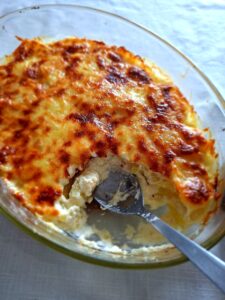 Top-down view of an oval glass baking dish with potato gratin with a golden and toasted cheese crust, with a portion already removed by a stainless steel spoon, revealing the creamy béchamel and potato interior.