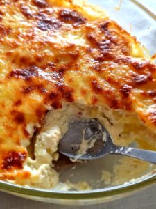 Close-up da travessa de vidro com gratinado de batata, mostrando em detalhe a crosta de queijo dourada e tostada e o interior branco e cremoso de molho branco e batata, com uma colher de inox a servir.