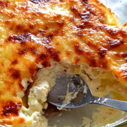 Close-up of the glass baking dish with potato gratin, showing in detail the golden and toasted cheese crust and the white creamy béchamel and potato interior, with a stainless steel spoon serving.
