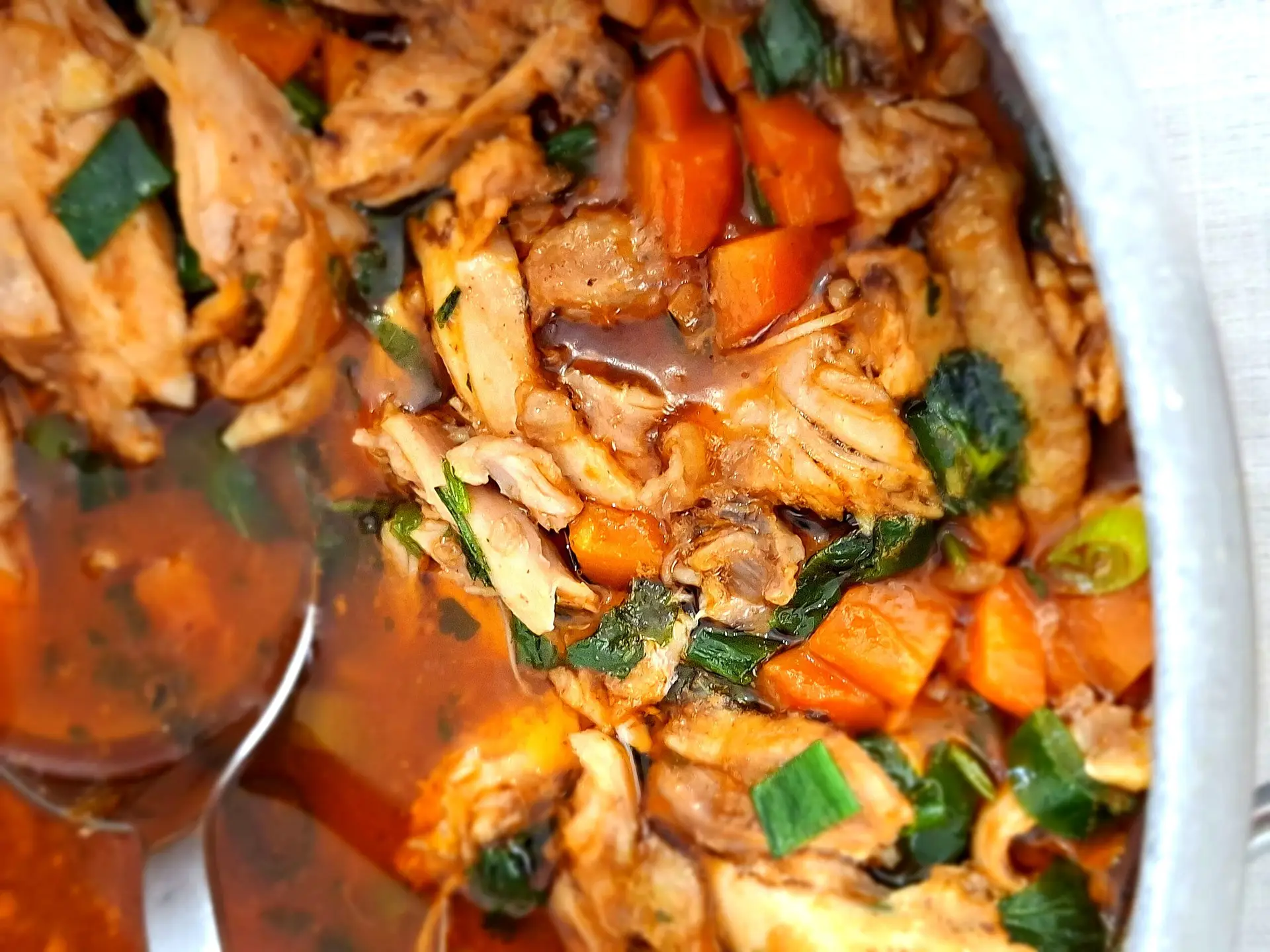 Close-up of a pot with shredded braised chicken in a dark orange sauce, with carrot pieces, green herbs and a stainless steel serving ladle.