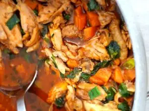Close-up de panela com frango estufado desfiado em molho alaranjado escuro, com pedaços de cenoura, ervas verdes e uma concha de servir em aço inoxidável.