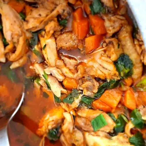 Close-up de panela com frango estufado desfiado em molho alaranjado escuro, com pedaços de cenoura, ervas verdes e uma concha de servir em aço inoxidável.