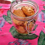 Frasco de vidro com fecho metálico tipo hermético, aberto, cheio de cookies artesanais com pepitas de chocolate, sobre toalha cor-de-rosa com estampado tropical de folhas e flamingos.