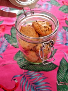 Open glass jar with a metal hermetic clasp, filled with artisan chocolate chip cookies, on a pink tablecloth with a tropical print of leaves and flamingos.