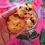 Mão aberta a segurar dois cookies artesanais diferentes: um com pepitas de chocolate escuro na parte superior e outro mais denso com pedaços de pistácio e frutos secos na parte inferior, junto ao frasco de vidro aberto.