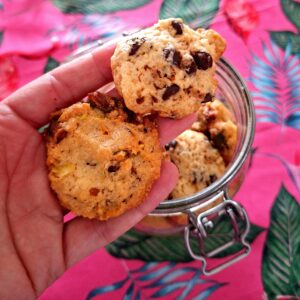 Open hand holding two different artisan cookies: one with dark chocolate chips on top and another denser one with pistachio pieces and dried fruit below, next to the open glass jar.