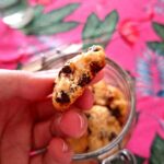 Mão a segurar um cookie artesanal com pepitas de chocolate junto ao bordo de um frasco de vidro aberto, com mais cookies visíveis no interior, sobre fundo rosa tropical.