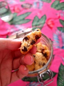 Hand holding a cookie broken in half, seen in profile, revealing a soft interior with dark chocolate chips, with the cookie jar blurred in the background.
