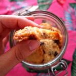 Mão a segurar metade de um cookie artesanal visto de perfil, com textura granulada e amanteigada visível no interior, sobre frasco com cookies e fundo rosa tropical desfocados.