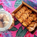 Vista aérea de frasco de vidro hermético com cookies e lata metálica aberta forrada a papel de manteiga com dois tipos de cookies artesanais — com chocolate e com pistácio — sobre toalha rosa com flamingos e folhas tropicais.