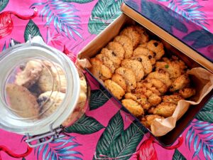 Aerial view of a hermetic glass jar with cookies and an open metal tin lined with parchment paper containing two types of artisan cookies — chocolate and pistachio — on a pink tablecloth with flamingos and tropical leaves.
