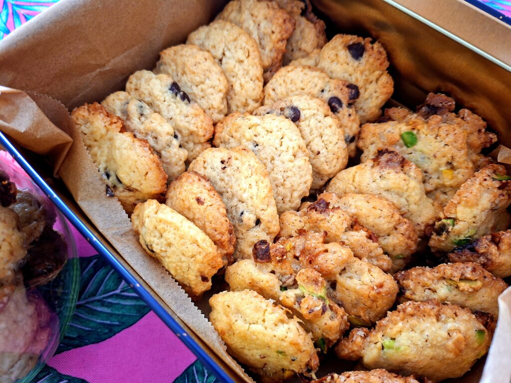 Close-up da lata metálica aberta com papel de manteiga, contendo dois tipos de cookies artesanais: com pepitas de chocolate escuro à direita e com pistácio e frutos secos à esquerda, com frasco de vidro parcialmente visível ao fundo.