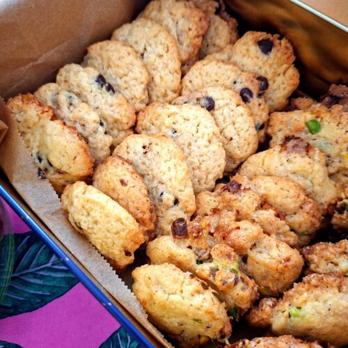 Close-up da lata metálica aberta com papel de manteiga, contendo dois tipos de cookies artesanais: com pepitas de chocolate escuro à direita e com pistácio e frutos secos à esquerda, com frasco de vidro parcialmente visível ao fundo.