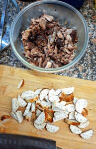 Aerial view of a granite worktop with a glass bowl of shredded pork and a bamboo chopping board with sliced German sausage cut into half-moons, with a chef's knife visible at the bottom.
