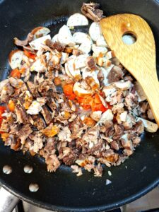 Aerial view of a dark non-stick wok with shredded pork, sliced German sausage and tomato pieces sautéing together, with an oak wooden spoon.