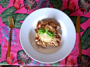 Aerial view of a deep white plate with spaghetti in a creamy sauce of shredded pork, German sausage and tomato, pasta nest at the centre with grated cheese and fresh basil leaves, on a pink tropical flamingo tablecloth with golden cutlery.