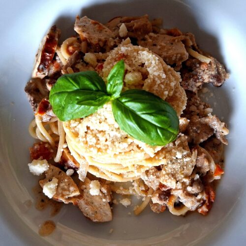 Close aerial view of a deep white plate with spaghetti in a creamy shredded pork and German sausage sauce, coiled pasta nest with grated cheese and two intensely green fresh basil leaves at the centre, on a pink tropical background.