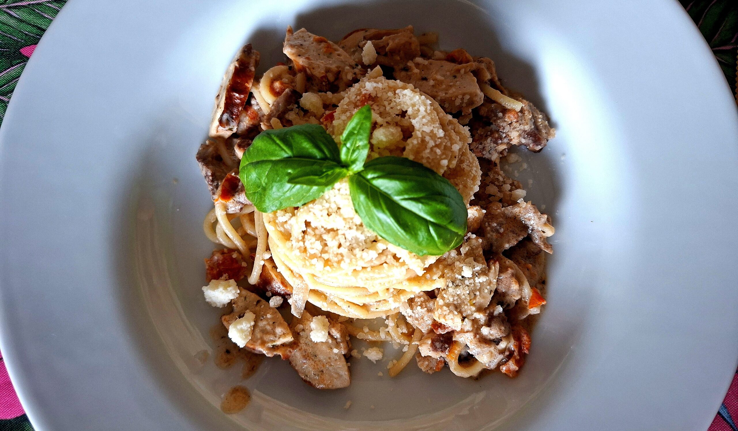 Close aerial view of a deep white plate with spaghetti in a creamy shredded pork and German sausage sauce, coiled pasta nest with grated cheese and two intensely green fresh basil leaves at the centre, on a pink tropical background.
