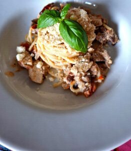 Slightly elevated frontal perspective of a deep white plate with spaghetti in a creamy shredded pork and German sausage sauce, pasta nest with grated cheese and basil leaves at the centre, on a pink flamingo tablecloth.