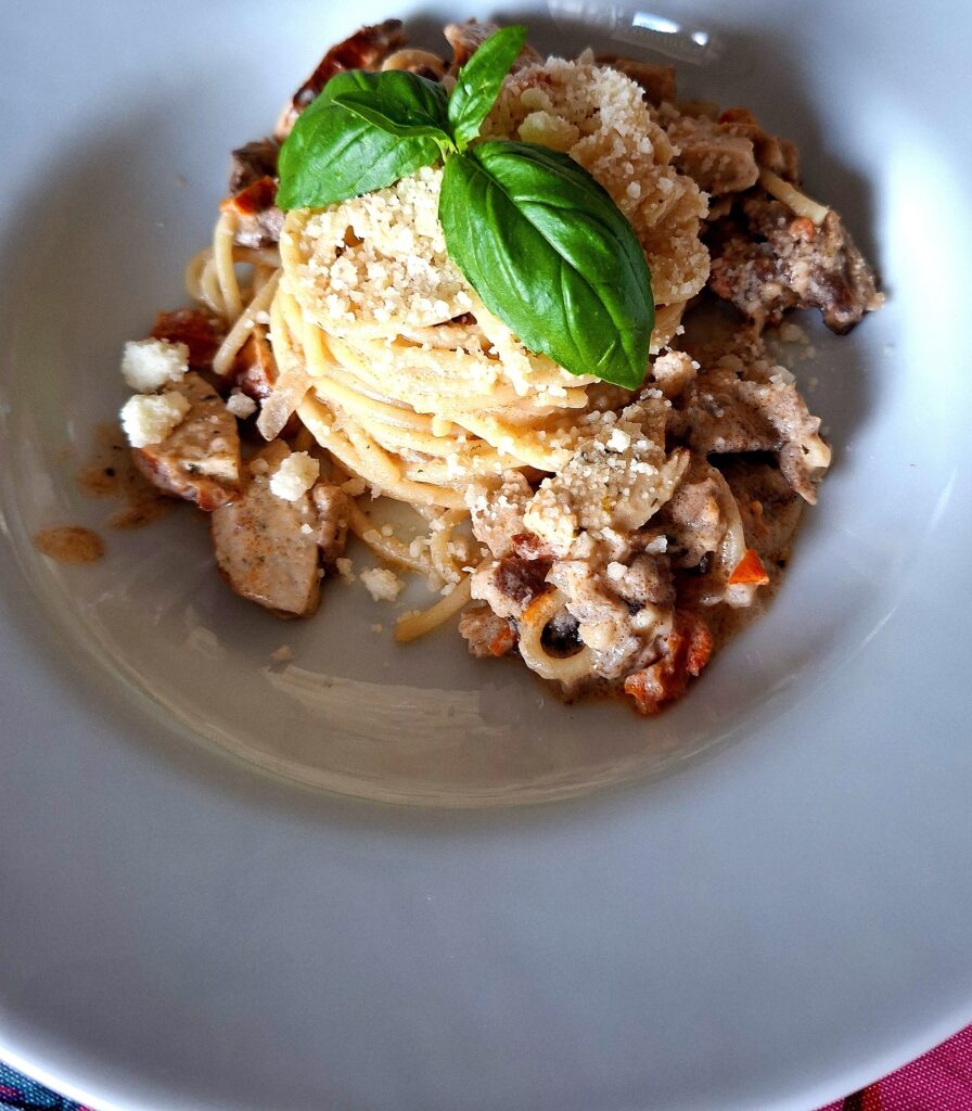 Slightly elevated frontal perspective of a deep white plate with spaghetti in a creamy shredded pork and German sausage sauce, pasta nest with grated cheese and basil leaves at the centre, on a pink flamingo tablecloth.