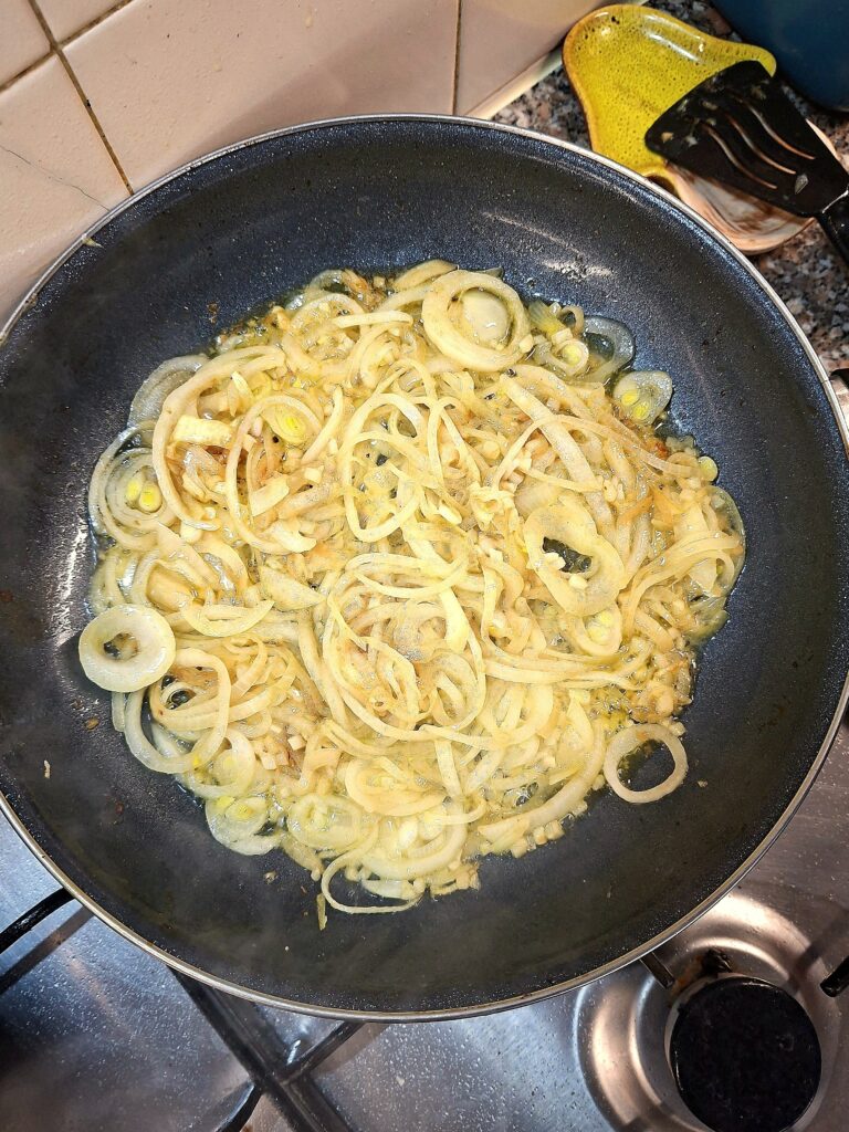 Sliced onions sautéing in a black frying pan, turning golden and translucent over the stovetop.