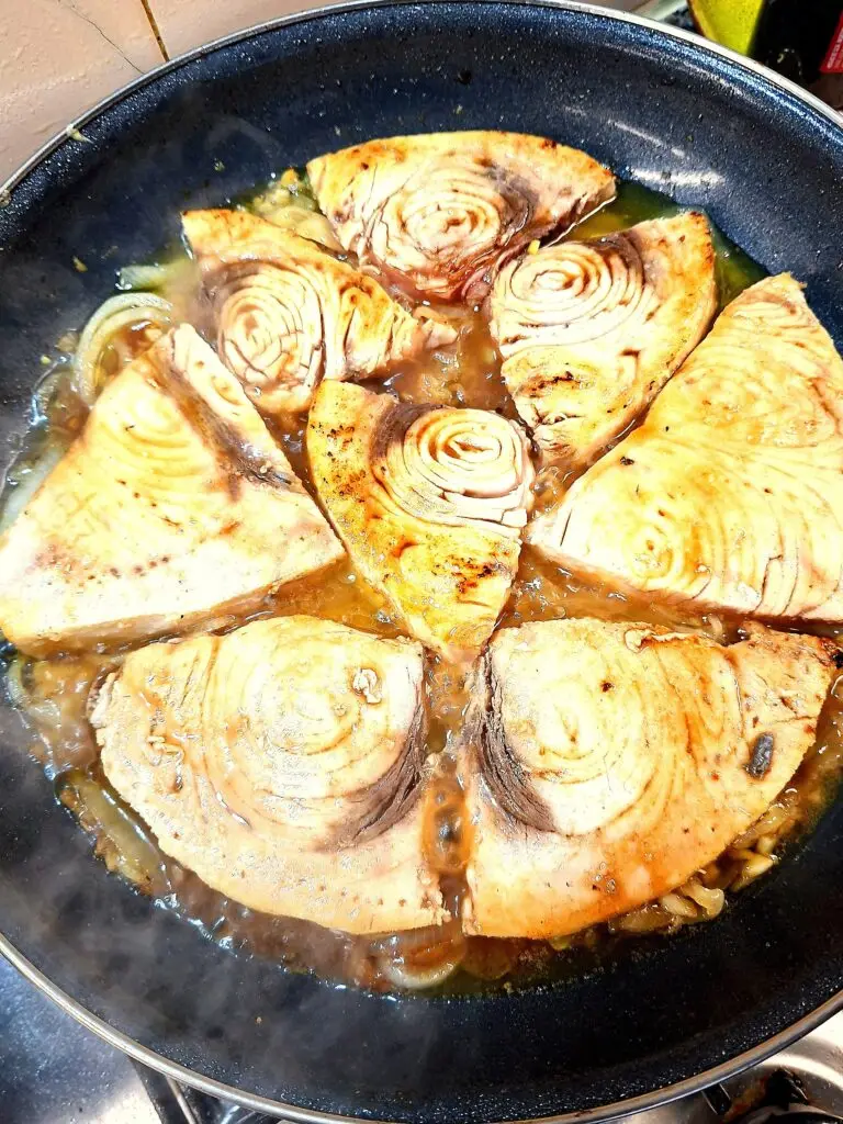 Postas de peixe a cozinhar num molho amarelo‑alaranjado dentro de uma frigideira larga, com especiarias visíveis no caldo.