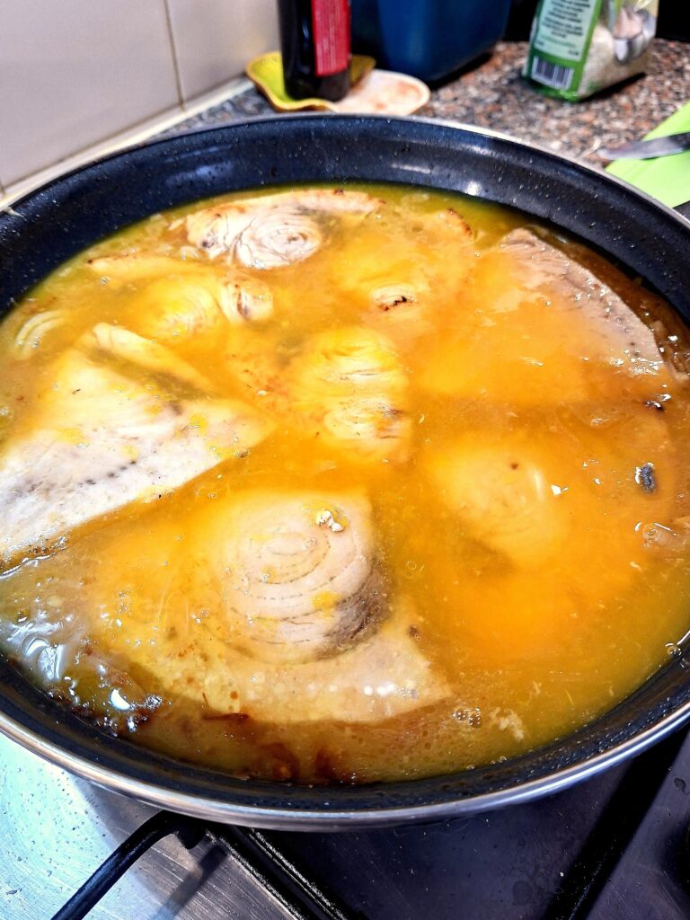 Postas de espadarte a cozinhar num molho espesso de cor laranja‑acastanhada, com pedaços de cebola e alho visíveis.